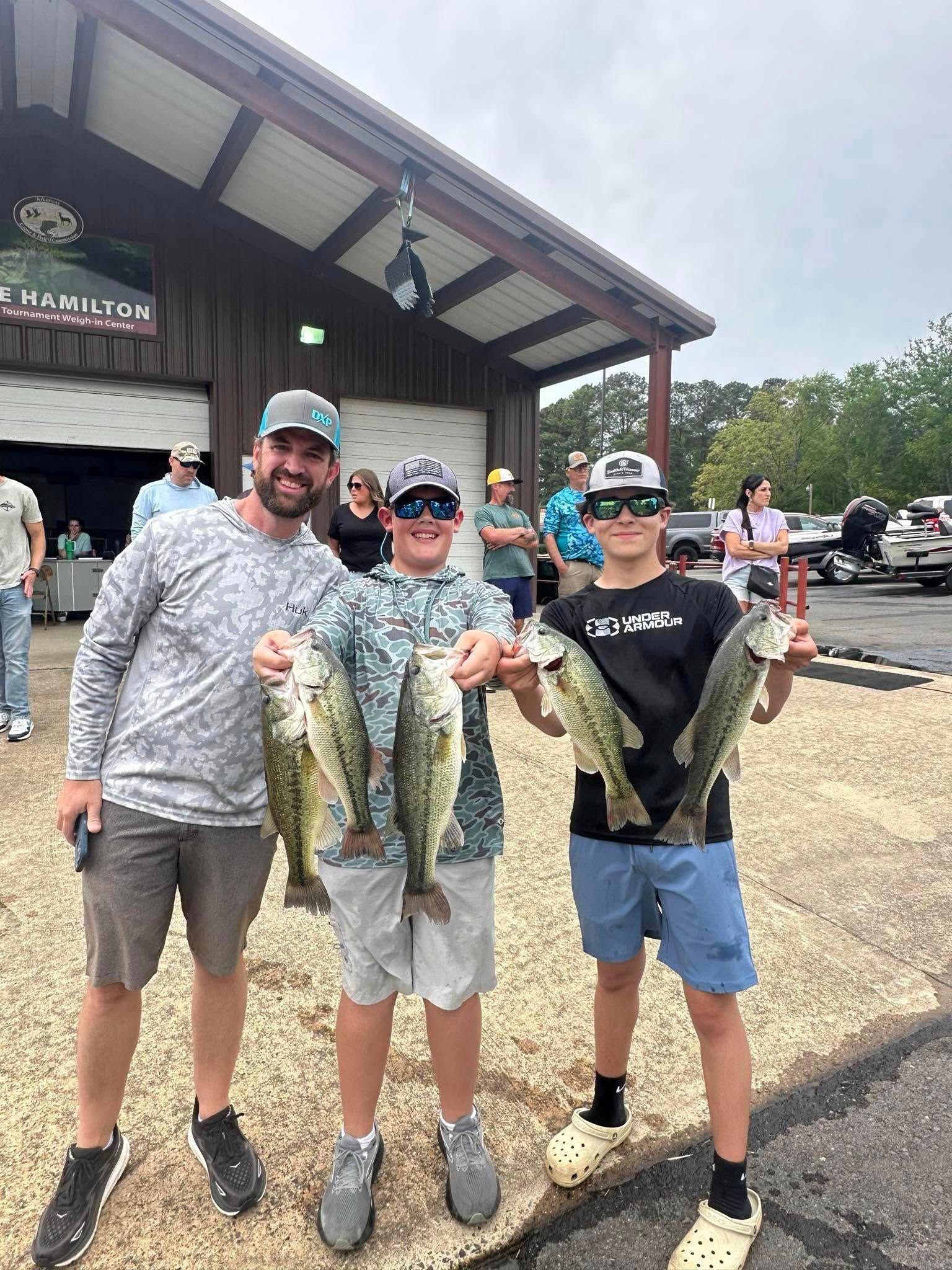Three people smiling and holding large fish outside a lakeside building.