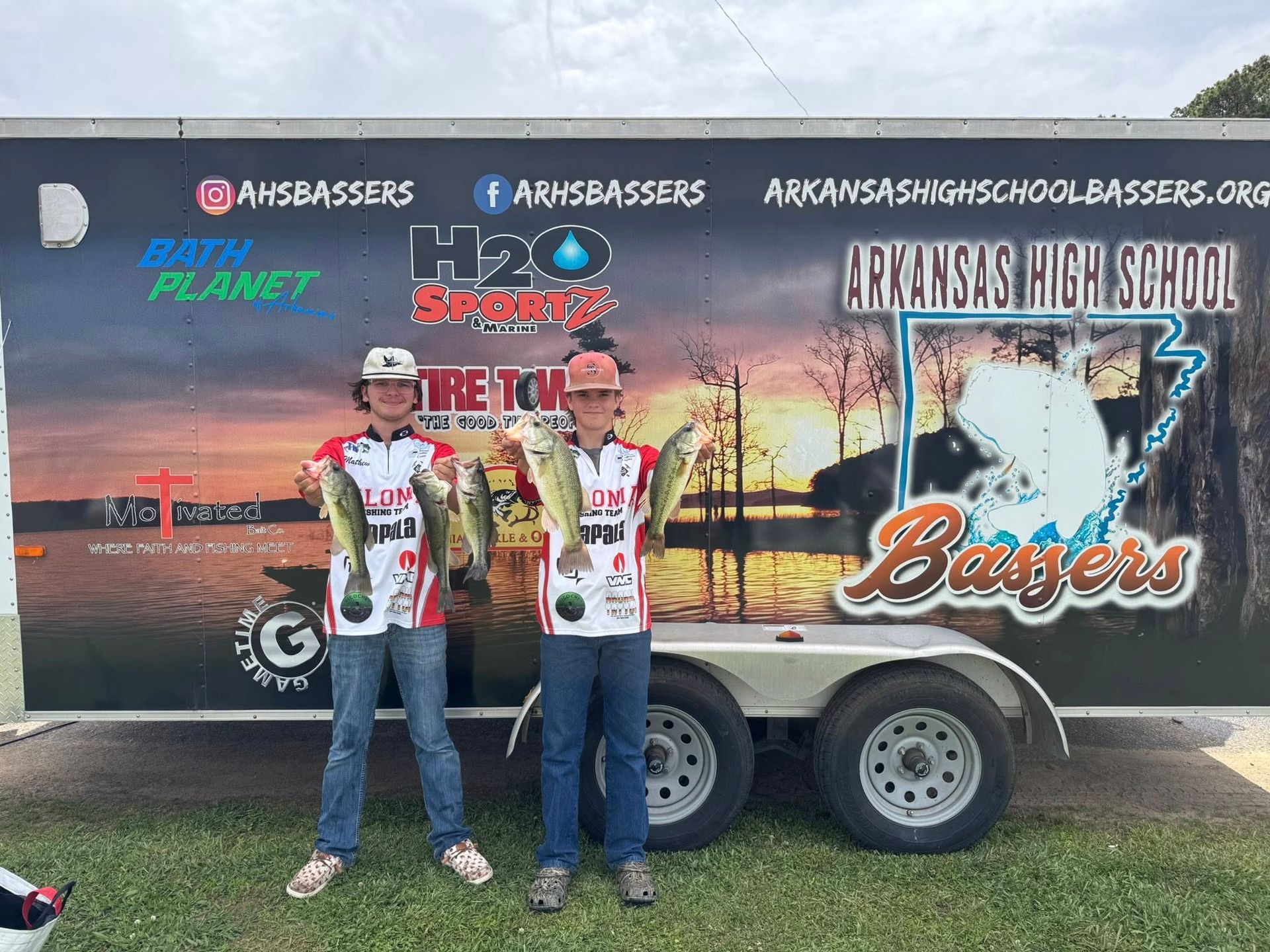 Two people in racing jerseys standing beside a trailer with Arkansas high school bass fishing graphics.