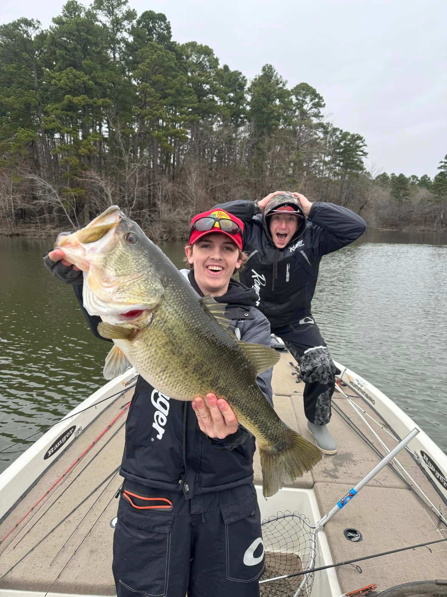 Two people on a boat hold a large bass in a wooded lake setting.