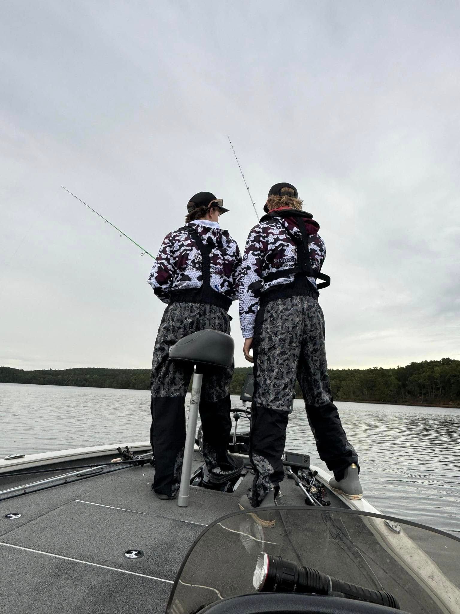 Two anglers in camouflage stand on a boat, fishing on a calm lake under a cloudy sky.