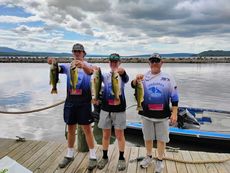 Three anglers holding large fish on a dock by the water under a cloudy sky