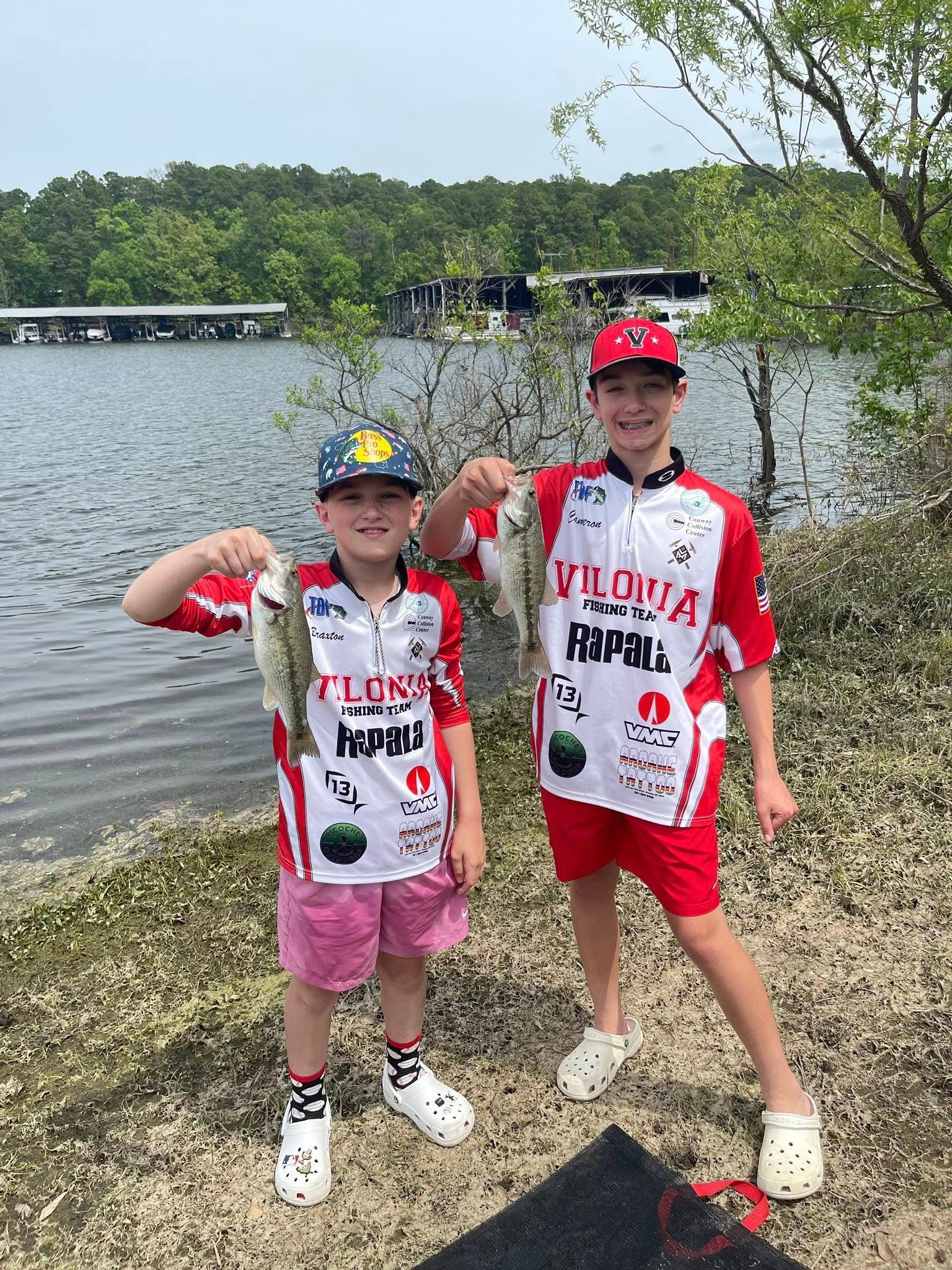 Two children in matching fishing jerseys hold up fish by a lakeshore.