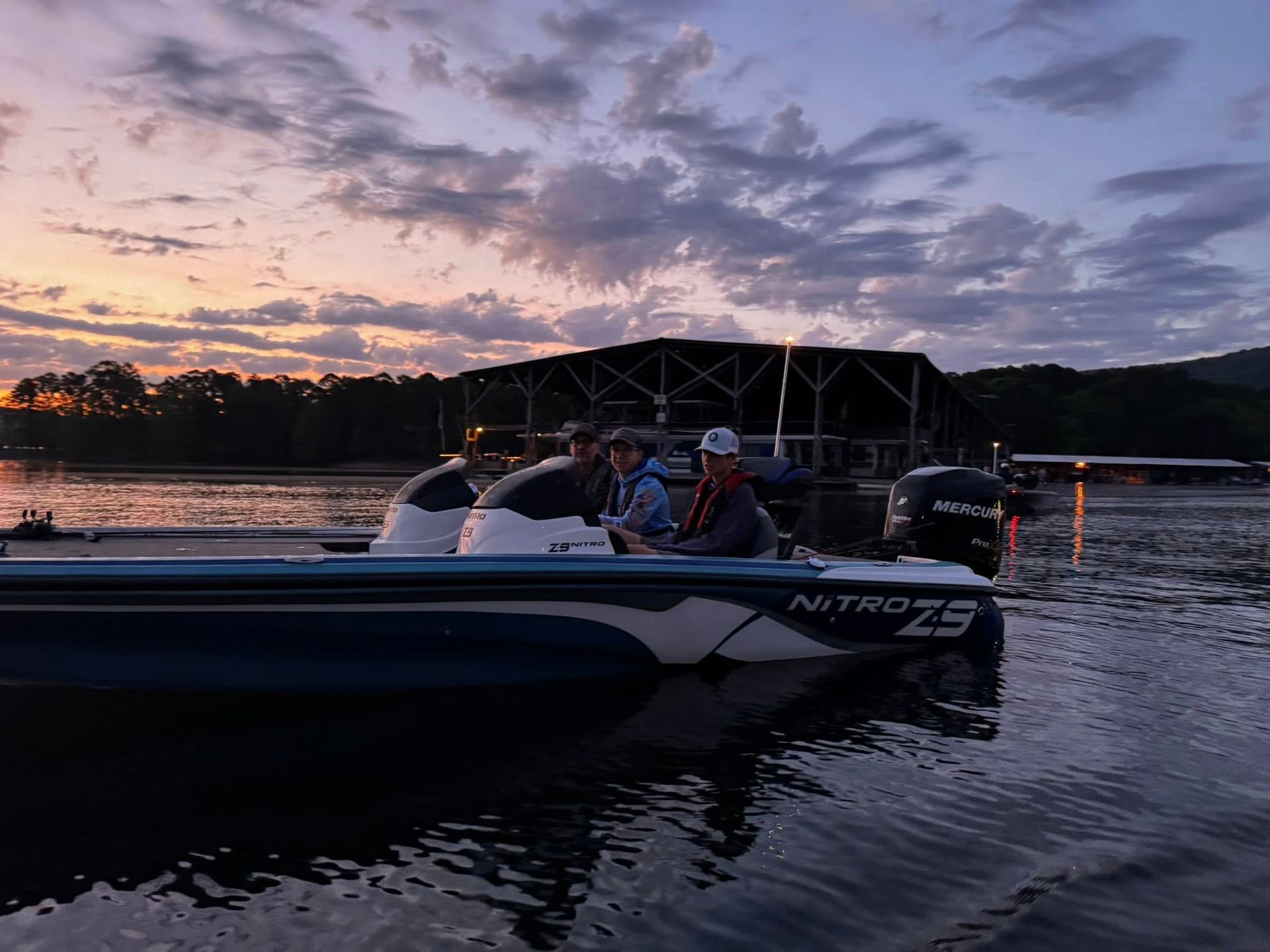 People in a white motorboat on a lake at sunset, with a dock and cloudy sky in the background.