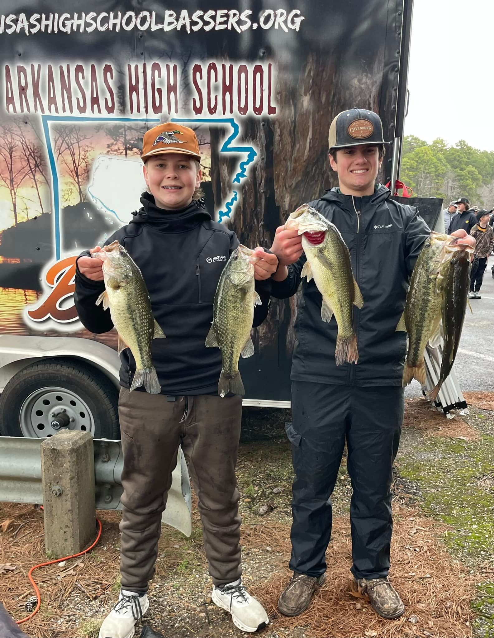 Two anglers holding large fish in front of a school bass fishing trailer