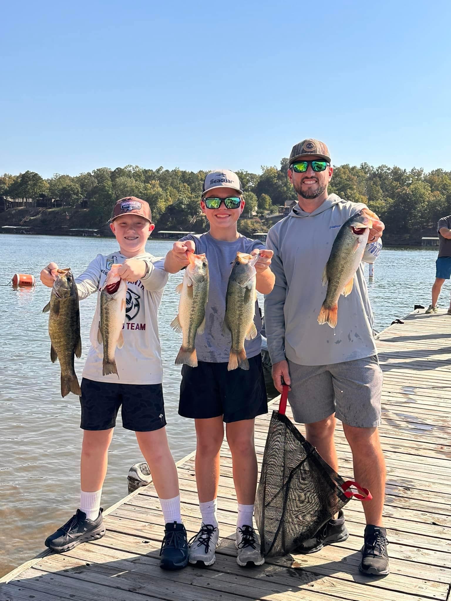 Three anglers on a lakeshore holding large fish, with a fishing net at their feet.