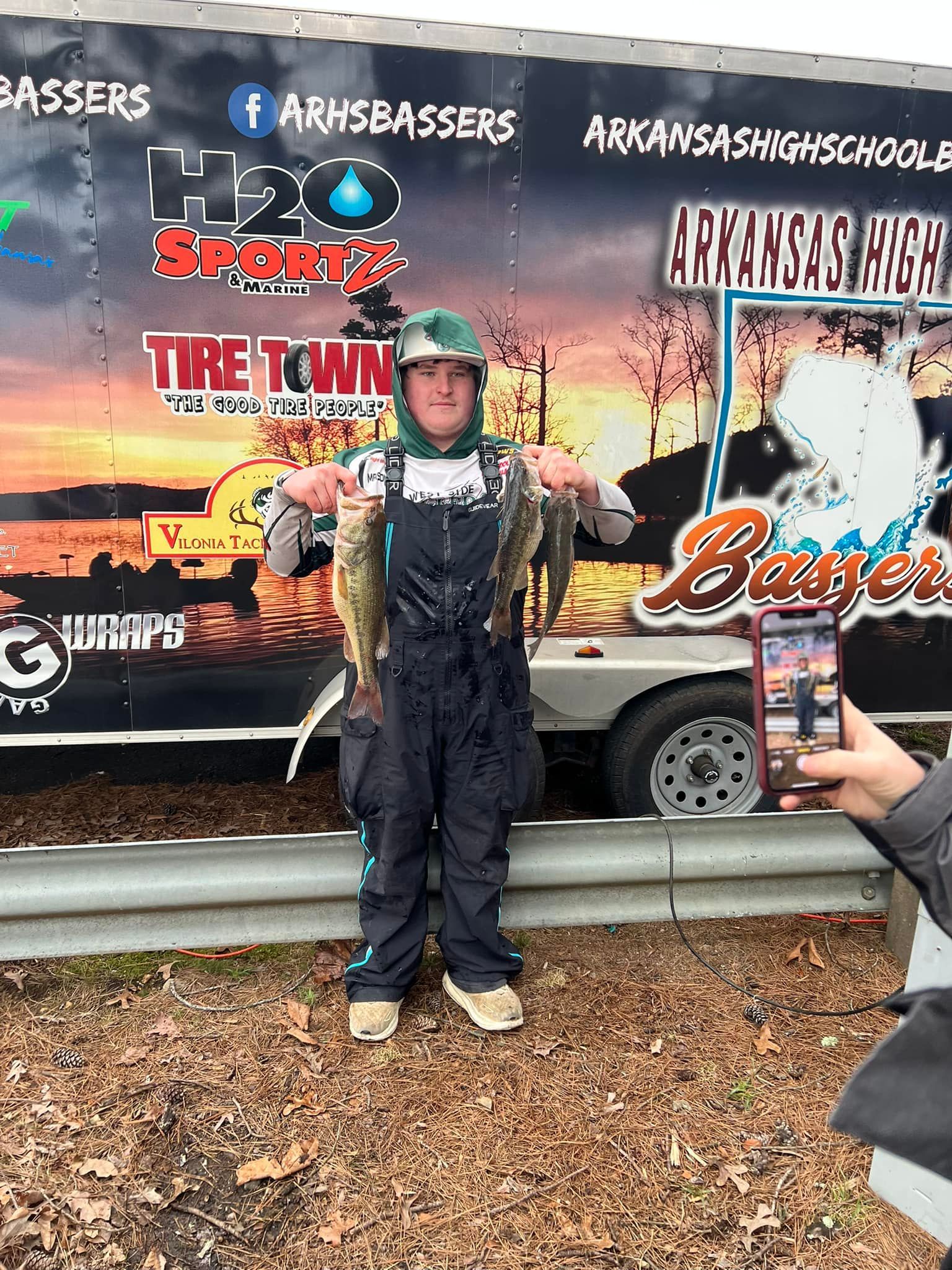 Child in helmet and protective gear stands in front of a Bass Pro Shops race trailer, posing for a photo.