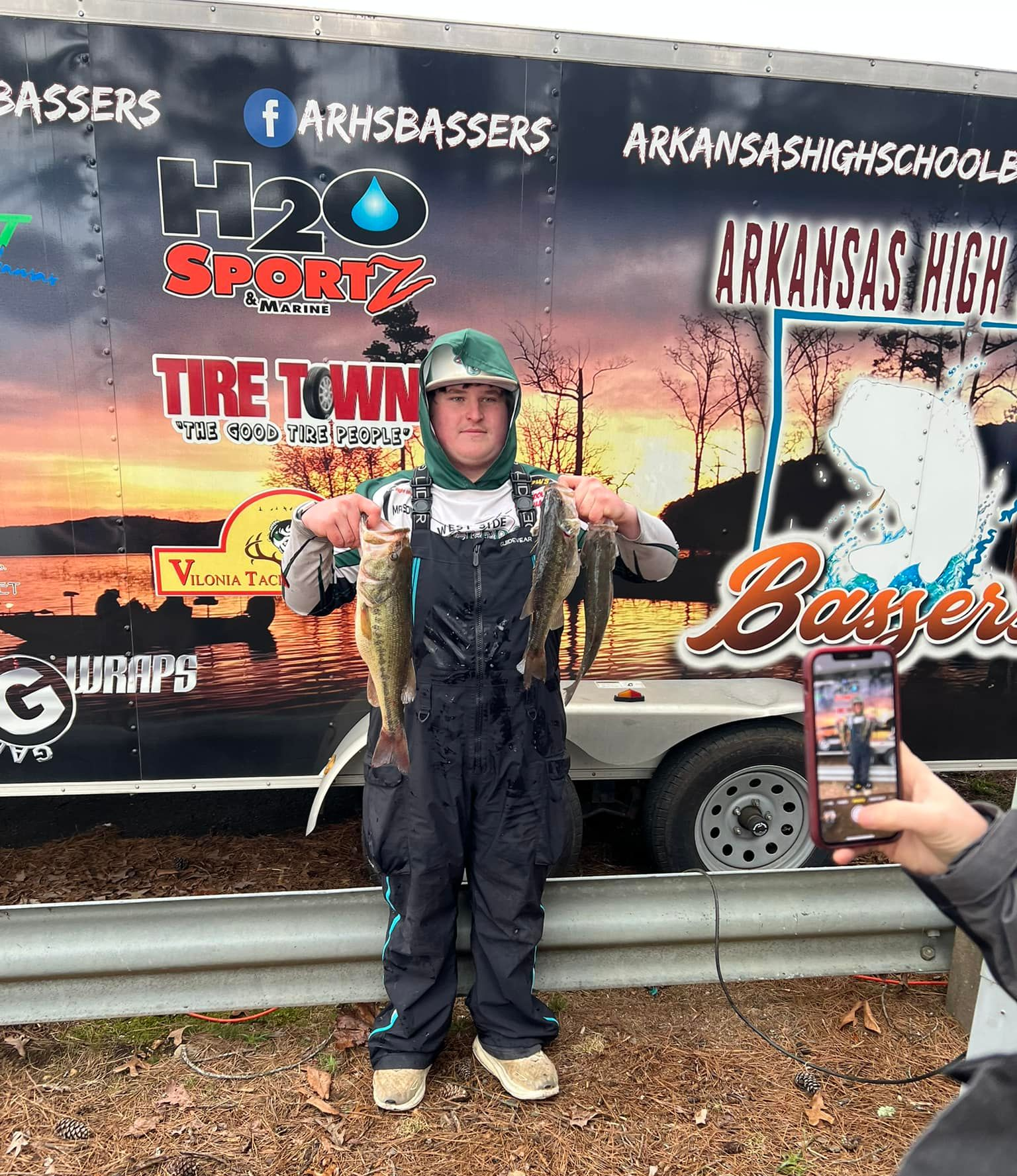Person in outdoor gear stands in front of a branded Arkansas Bassmasters trailer, holding a fish trophy.