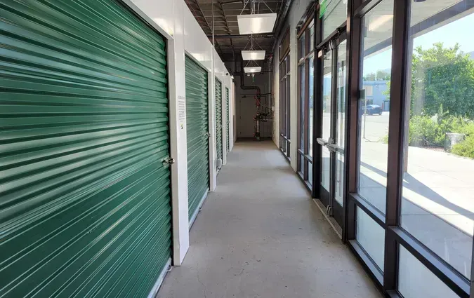 A hallway with green roll-up storage unit doors on the left and glass doors and windows on the right.