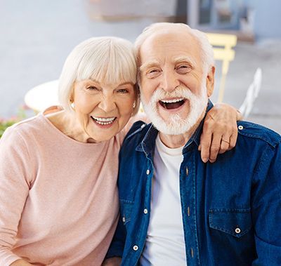 Smiling couple with arm around each other, outdoors. Man in denim shirt, woman in pink.