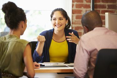 Woman in yellow top gestures, speaking to a couple at a desk. Brick wall, bright lighting.