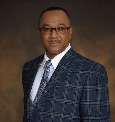 Man in blue plaid suit, glasses, and paisley tie, posed against a brown background.