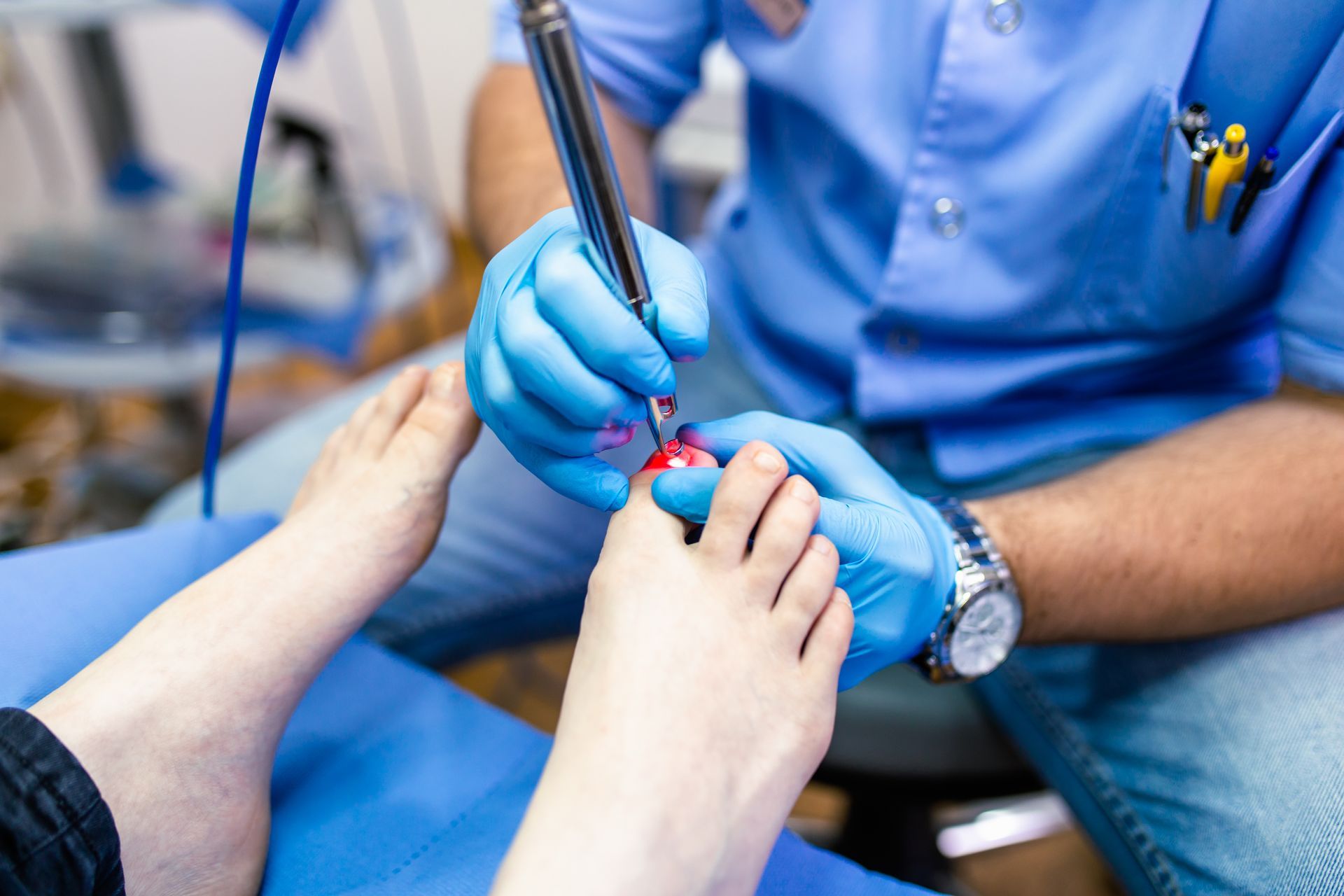 Podiatrist using a tool on a patient's toe, wearing blue gloves and scrubs, in a medical setting.
