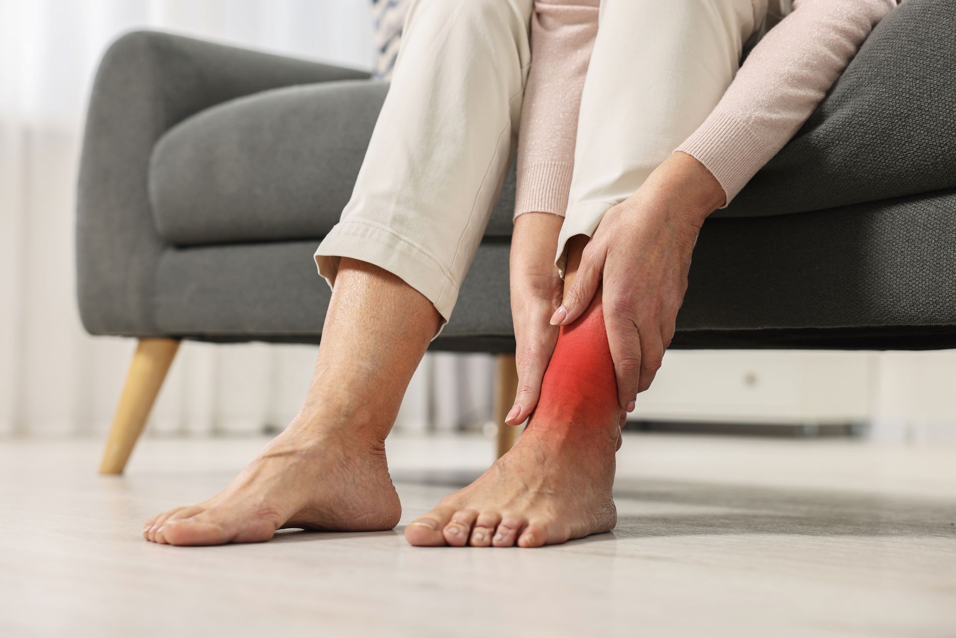 Woman sitting on a gray couch, holding ankle in pain, red discoloration indicating injury.