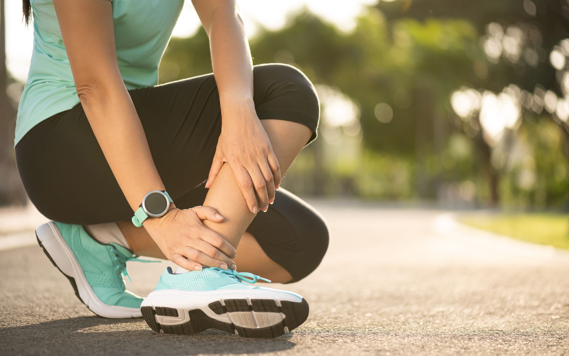 Person holding ankle, possibly injured after running outside on a paved path, wearing athletic wear and watch.