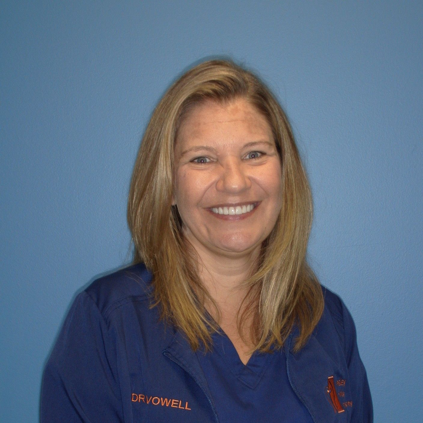 Woman with blonde hair in blue scrubs smiles at the camera, blue background.