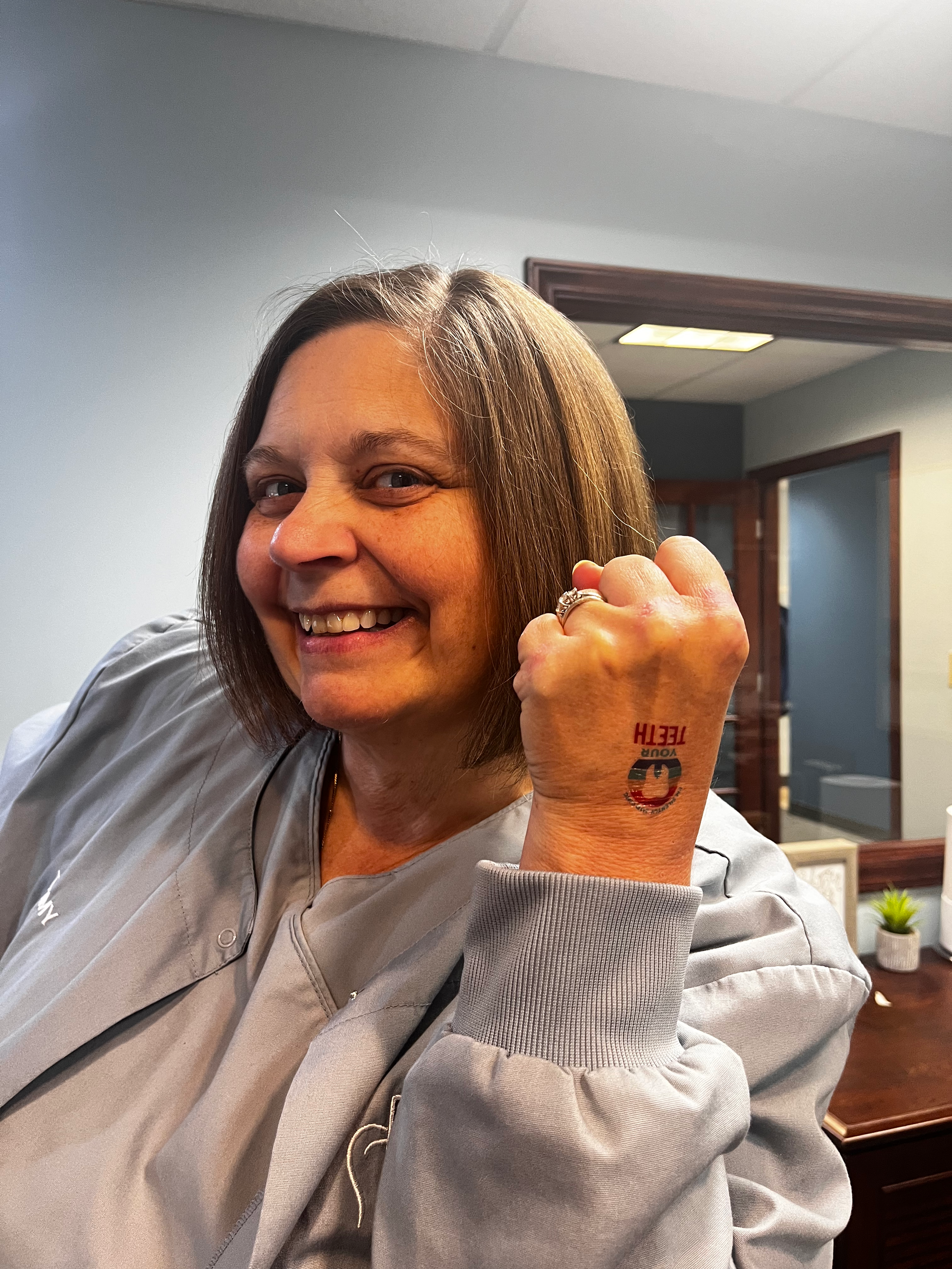Woman with a tattoo on her wrist, smiling. She's wearing a grey jacket, indoors.