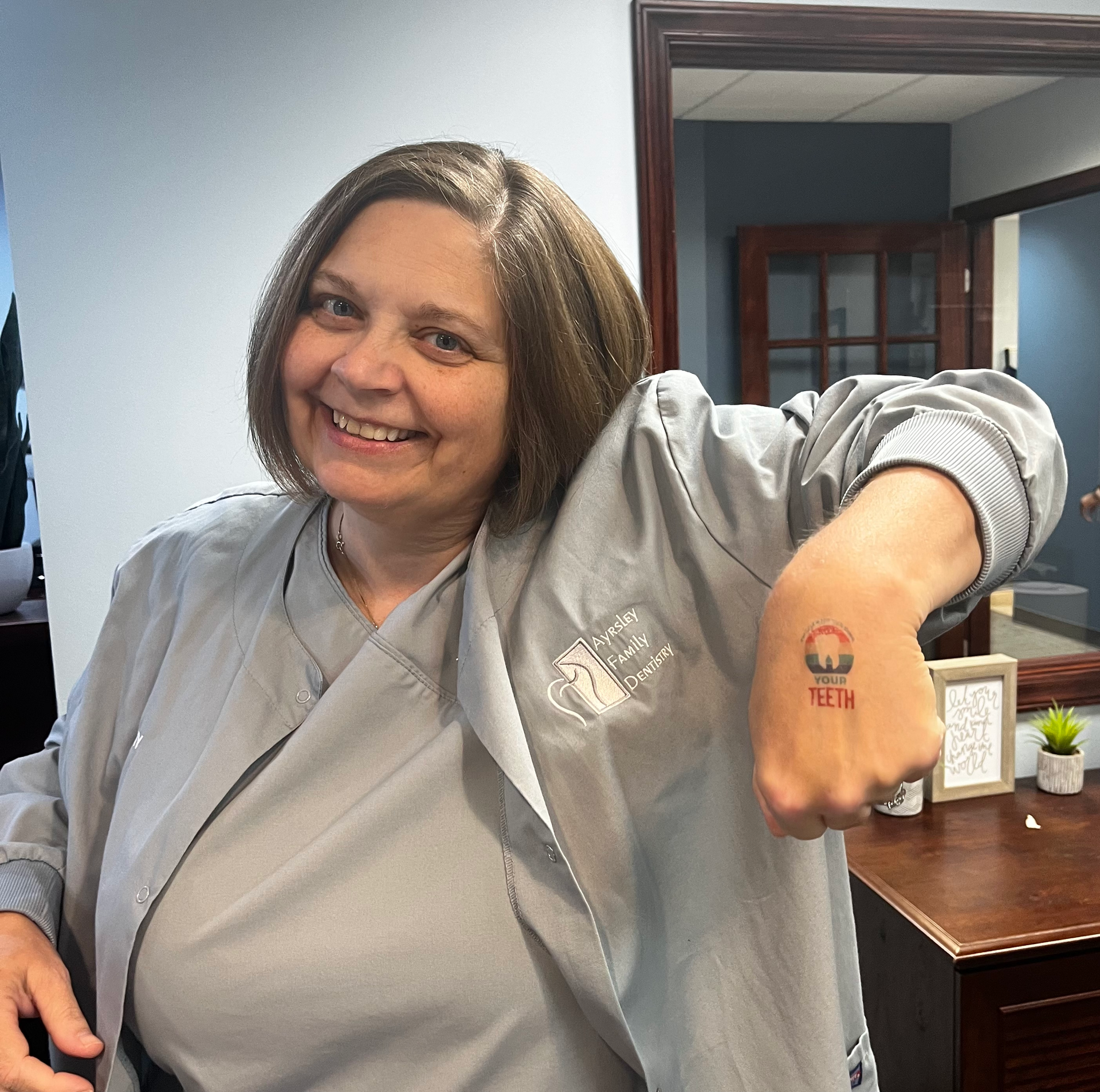 Woman with a gray jacket smiles, showing a fist tattoo. Indoors with a desk and doorway in the background.