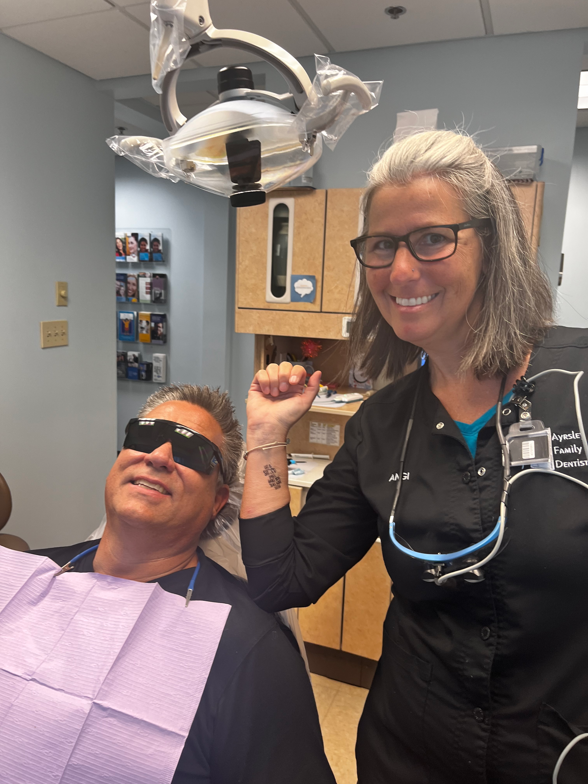 Dentist and patient. Smiling dentist with fist raised, patient in chair wearing shades. Dental office setting.