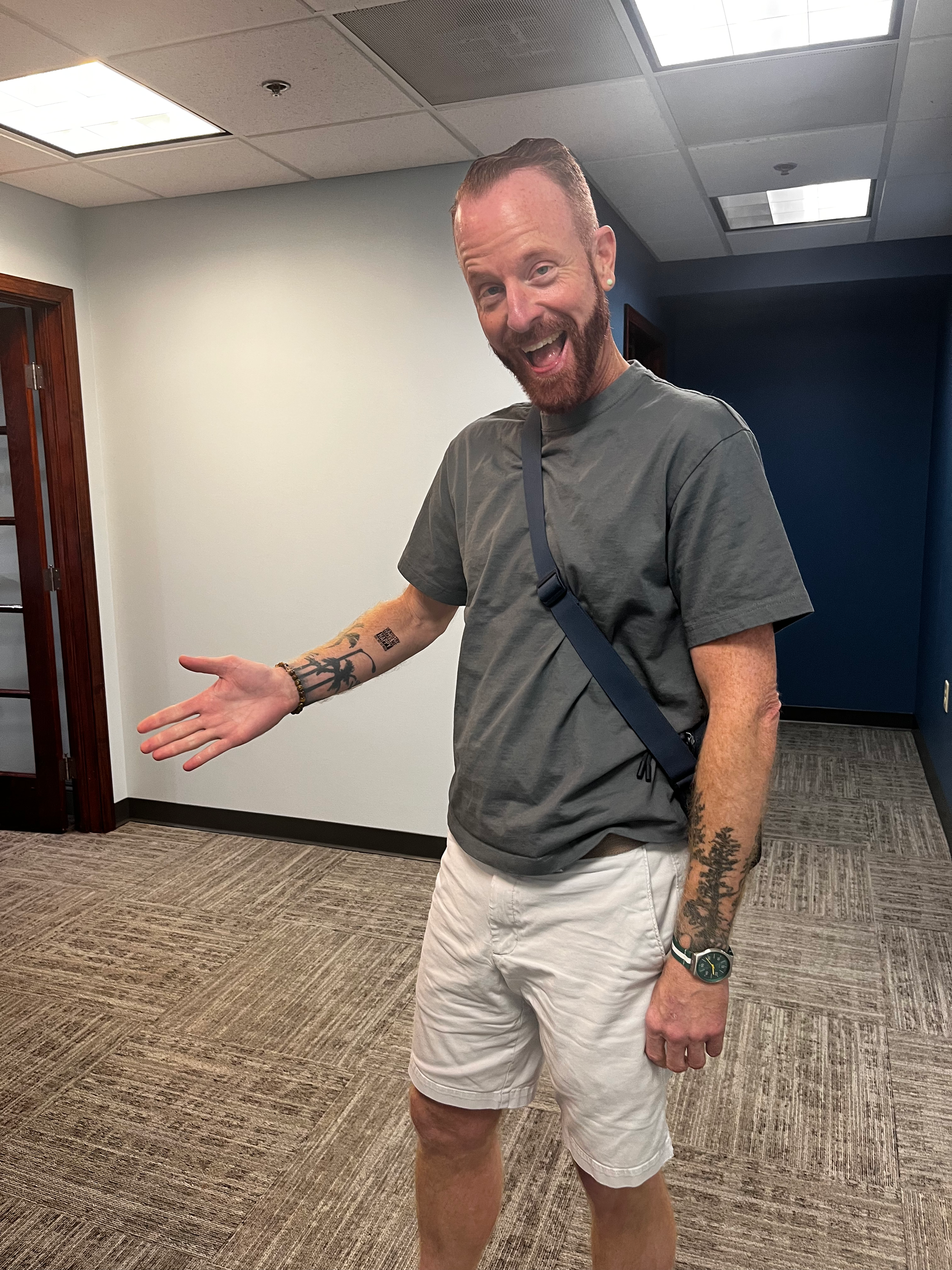 Man with tattoos and grey shirt smiles and gestures towards the left. Standing in an office hallway.