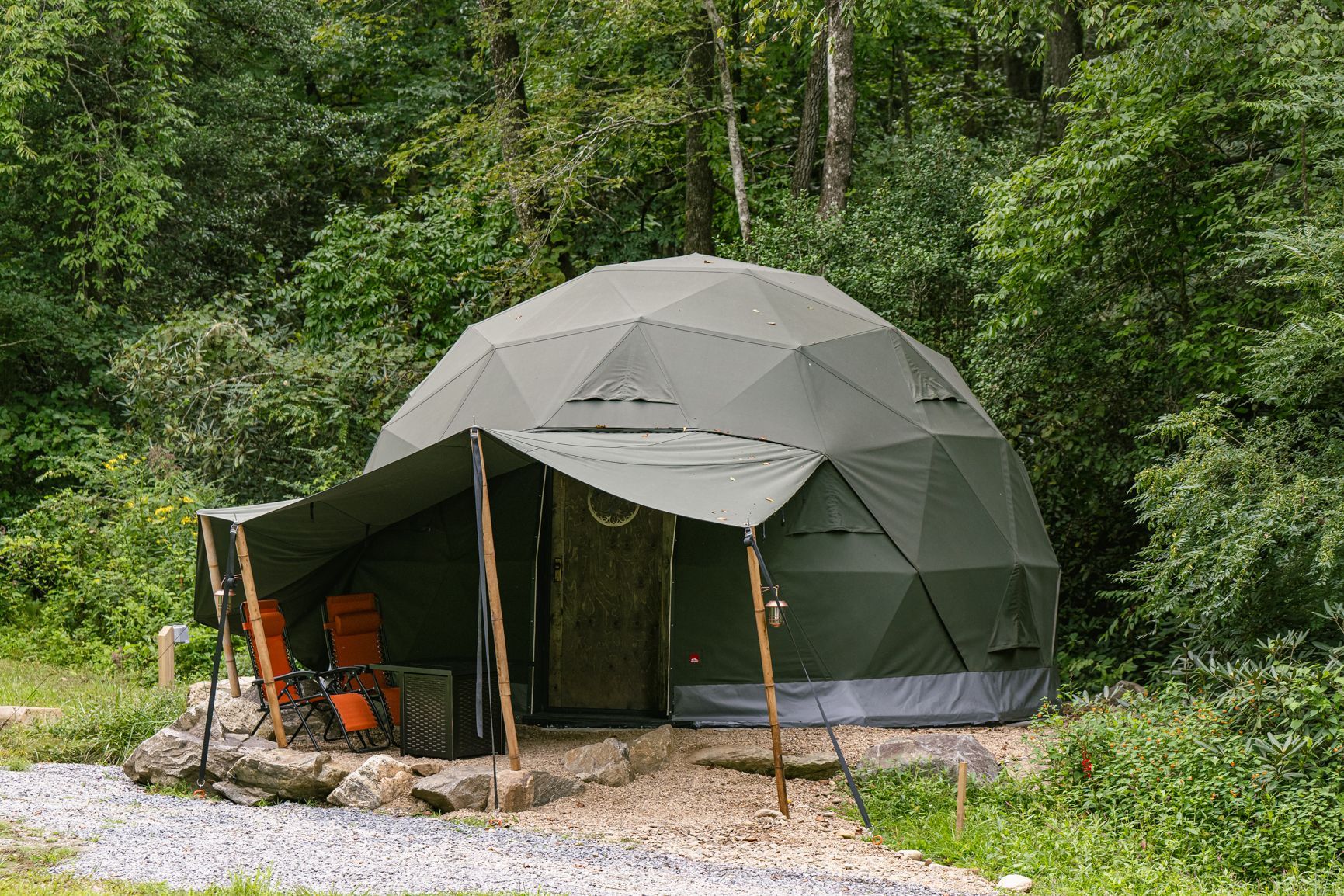 Green geodesic dome glamping tent with awning, surrounded by trees and gravel. Two orange chairs on the porch.