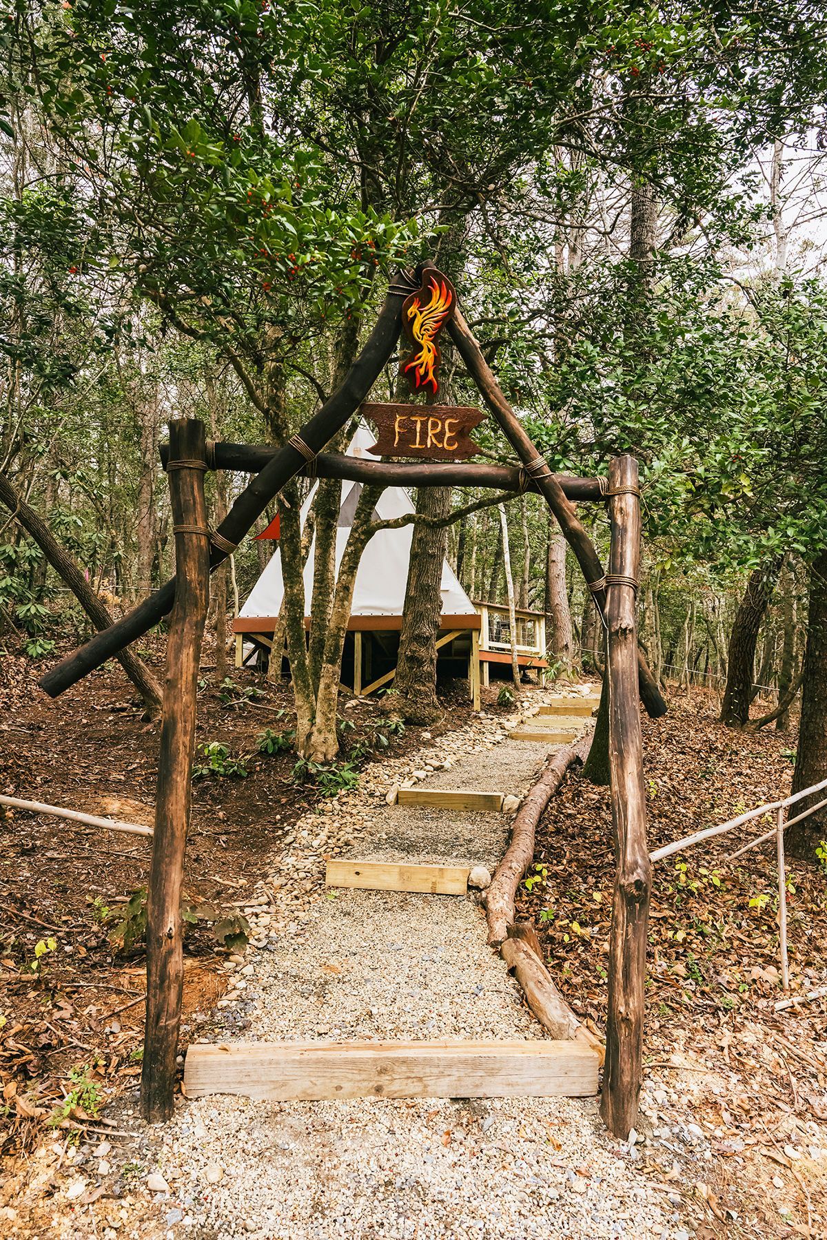Rustic wooden archway leads to a teepee structure in a wooded area. 