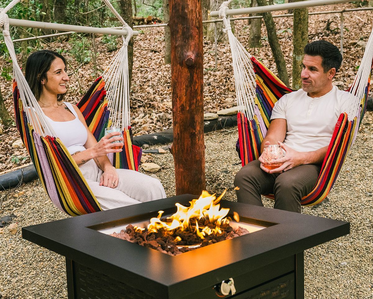 Couple relaxing in hammocks by a fire pit in a wooded area.