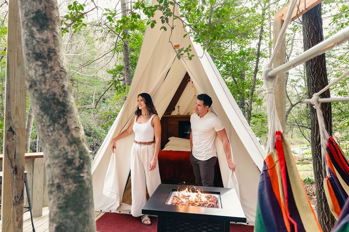 Couple exits a canvas tent yurt. A fire pit sits in front of the open tent with trees and a hammock nearby.