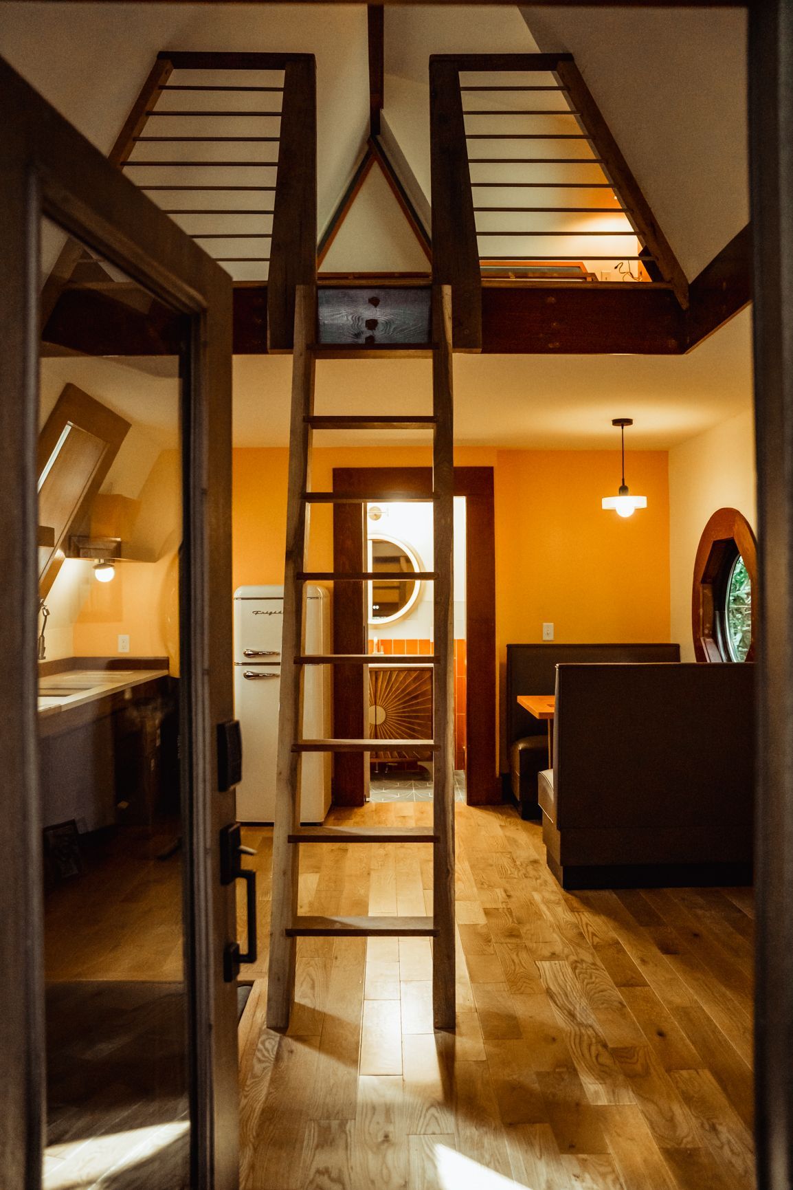 Interior view of a small cabin with a ladder leading to a loft. Orange walls, wood floor, and a glimpse of a kitchen.