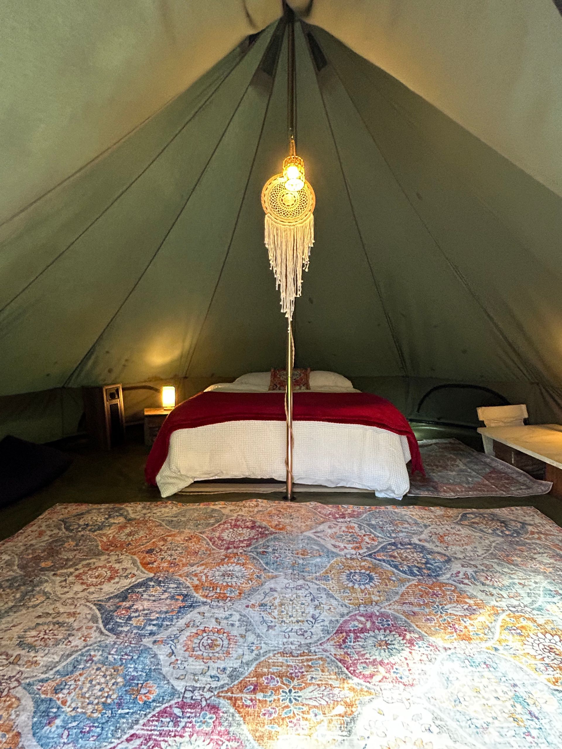Inside a canvas tent: bed with red blanket, decorative rug, hanging light, and side tables.
