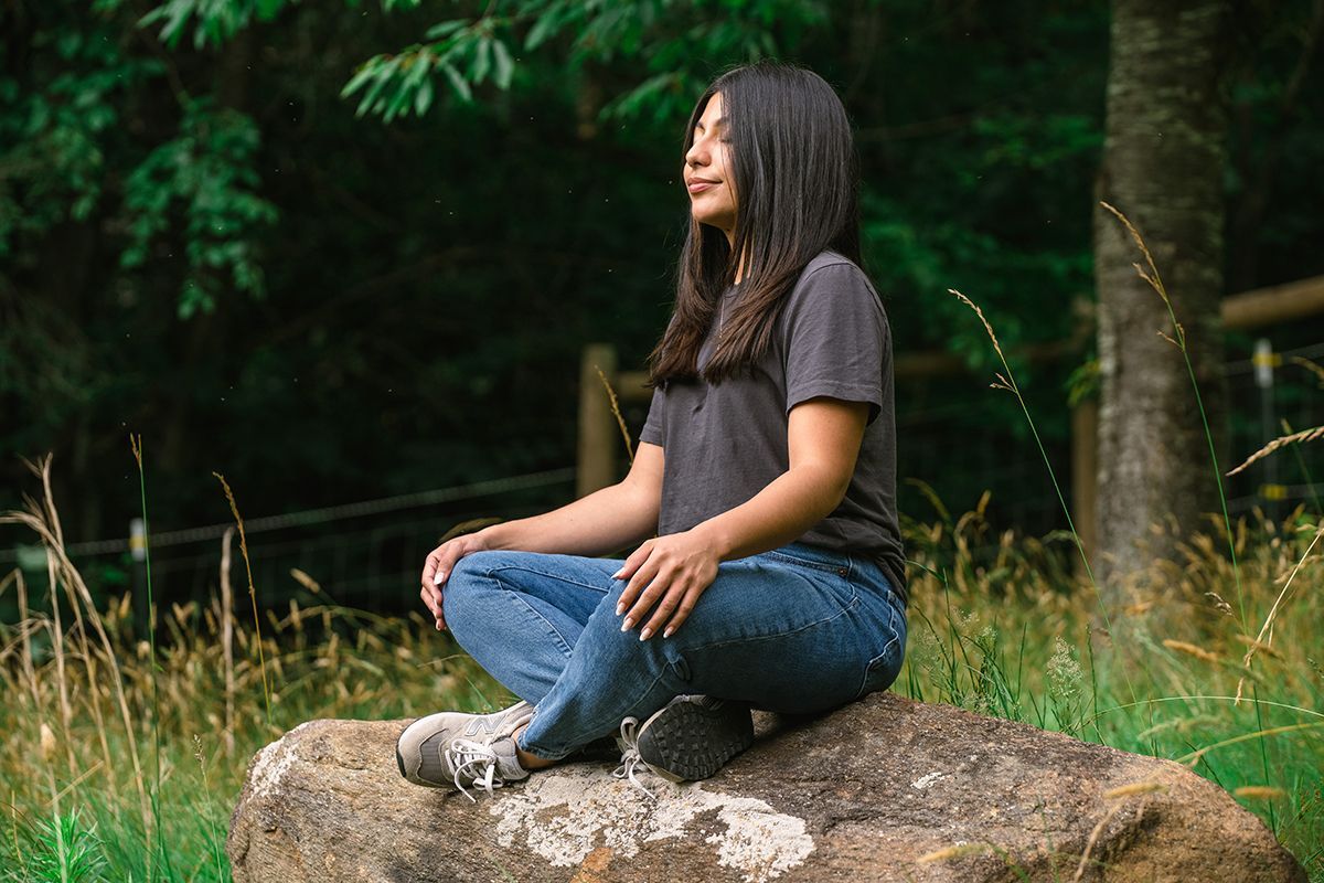 A woman is sitting in a lotus position on a rock in the woods.