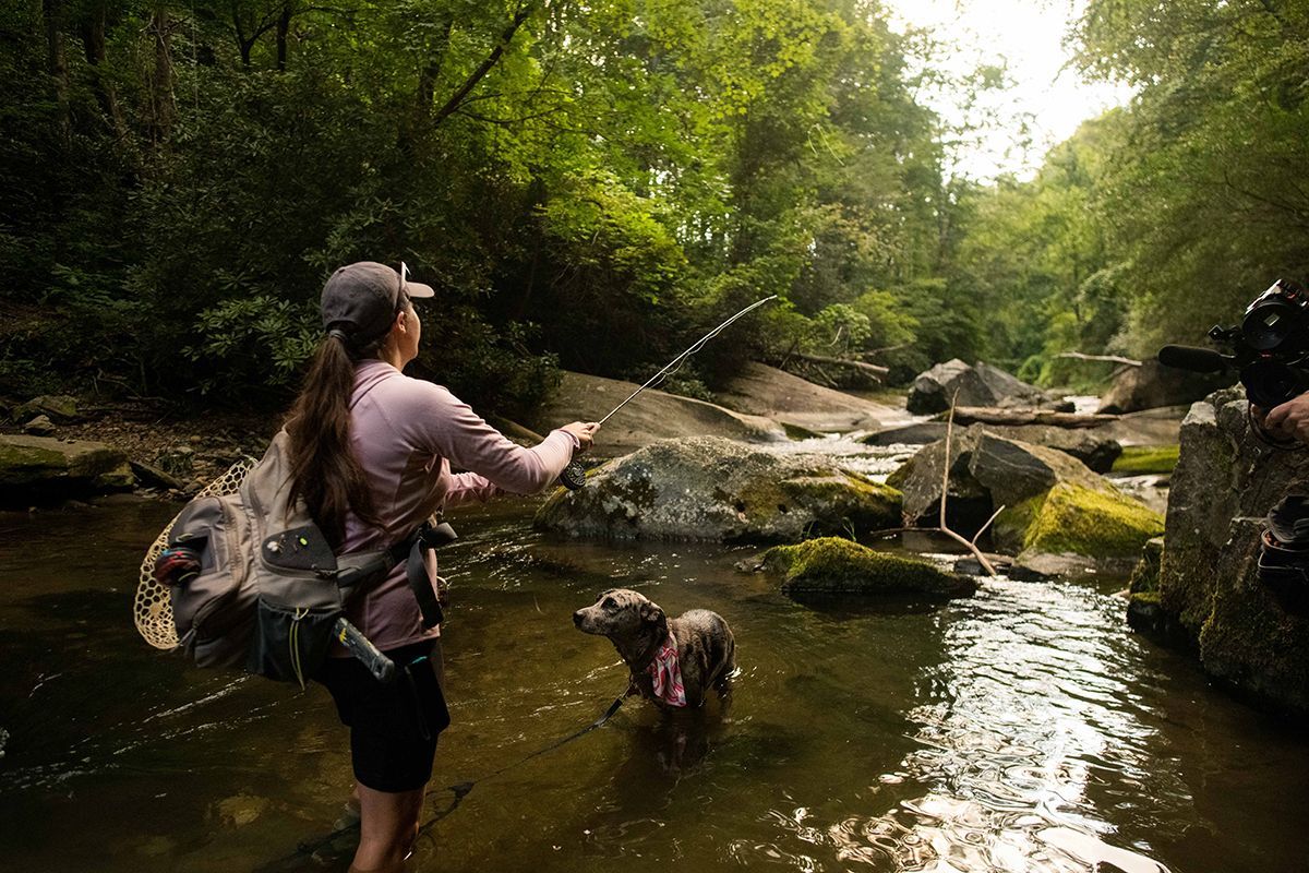 A woman fly fishes with her dog in a creek.