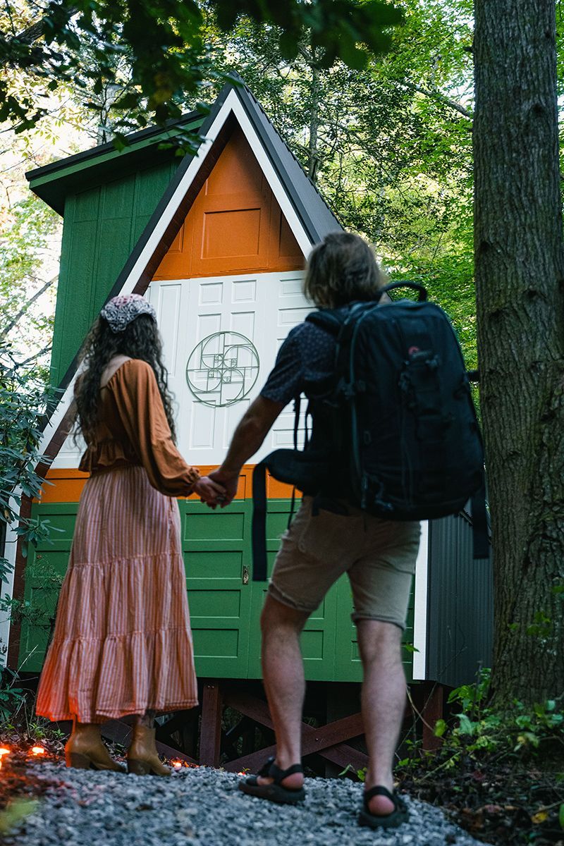 Couple holding hands, facing a colorful wooden creator cabin in a forest. One wears a backpack.
