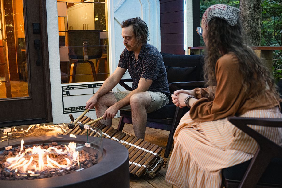 Man playing a marimba near a fire pit with a woman seated beside him outdoors.