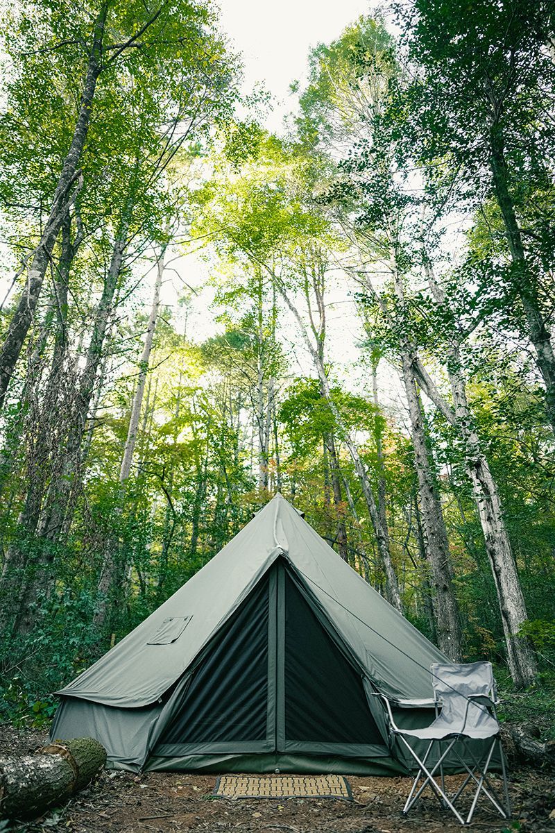Canvas tent in a forest, chair nearby, tall green trees above.