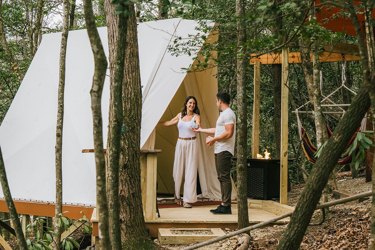 Couple standing on the porch of a canvas tent in a wooded area, smiling.
