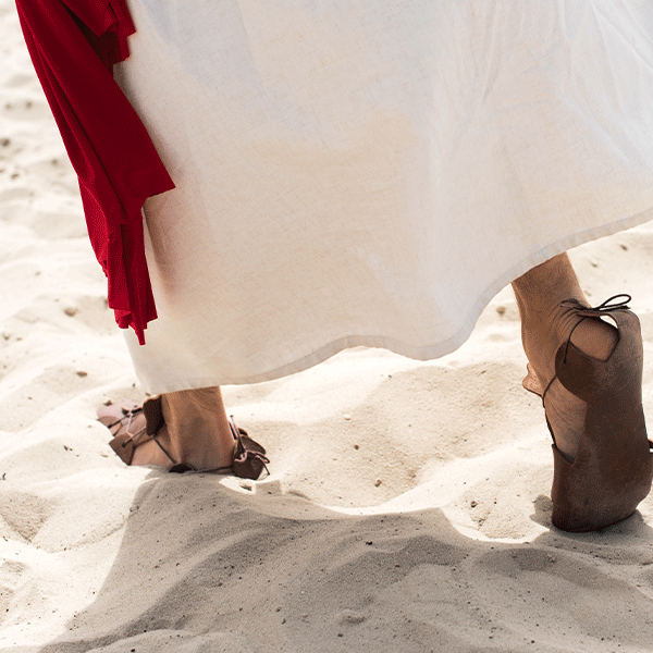 Person in white robe and sandals walking on sand, red cloth visible.
