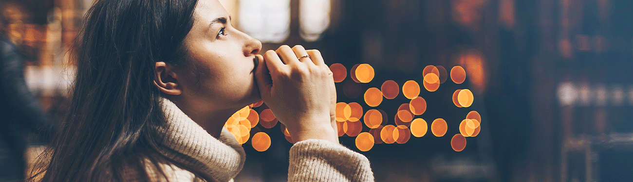 A woman prays inside a dark church. Candlelight glows in the background.