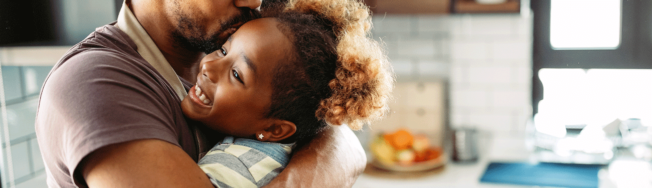 A person hugs a smiling child in a kitchen. The child is wearing a blue and white patterned shirt.