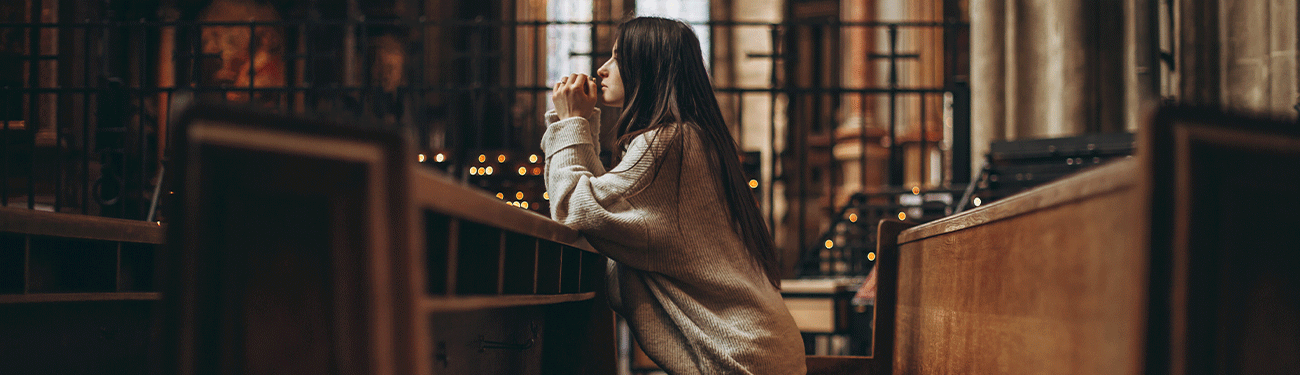 A woman with long dark hair, wearing a knitted sweater, kneels in prayer at a wooden pew inside a dimly lit cathedral.