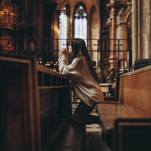 A person kneels in prayer in a dimly lit, wooden-pew church with stained glass windows in the background.