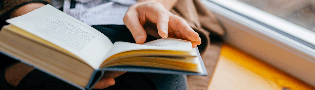 A person holds an open book, turning a page, while sitting near a window with yellow light.