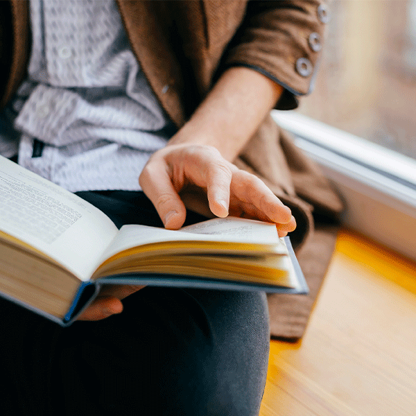 A person in a brown jacket sits by a window, turning the page of an open book held in their lap.
