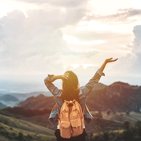 Woman with backpack raises arms to sky atop mountain.
