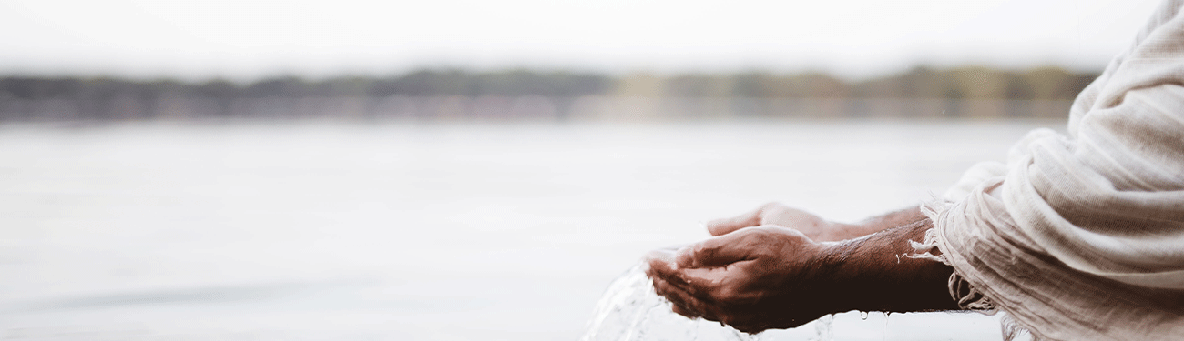 Hands cupping water near a body of water; light beige clothing in the frame.