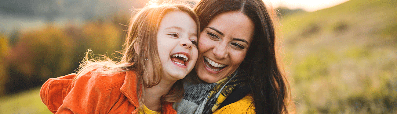 A woman and a girl laughing together outdoors in front of a blurred background.
