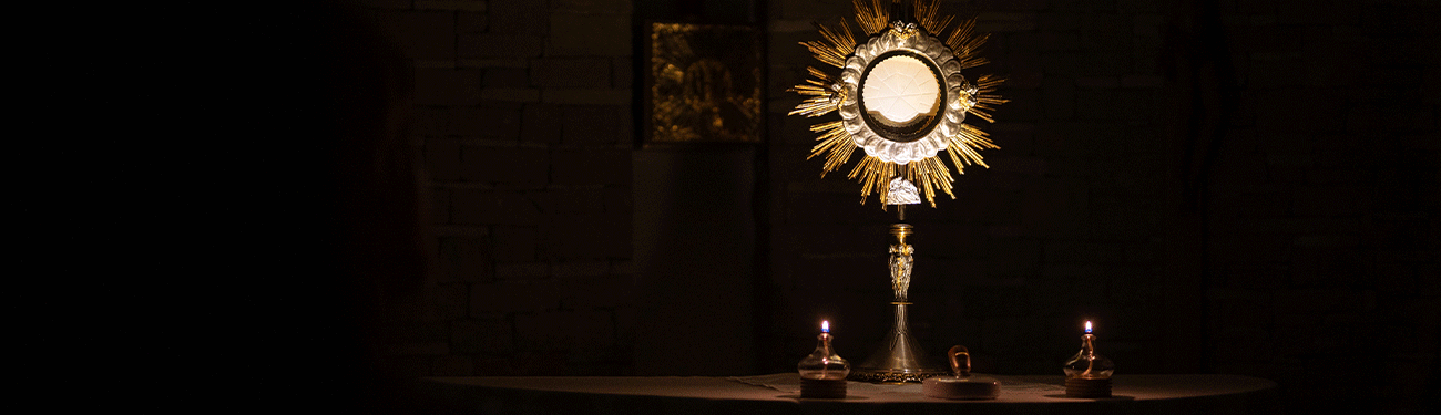 A gold monstrance containing the Eucharist stands illuminated on an altar in a dark church, flanked by two candles.