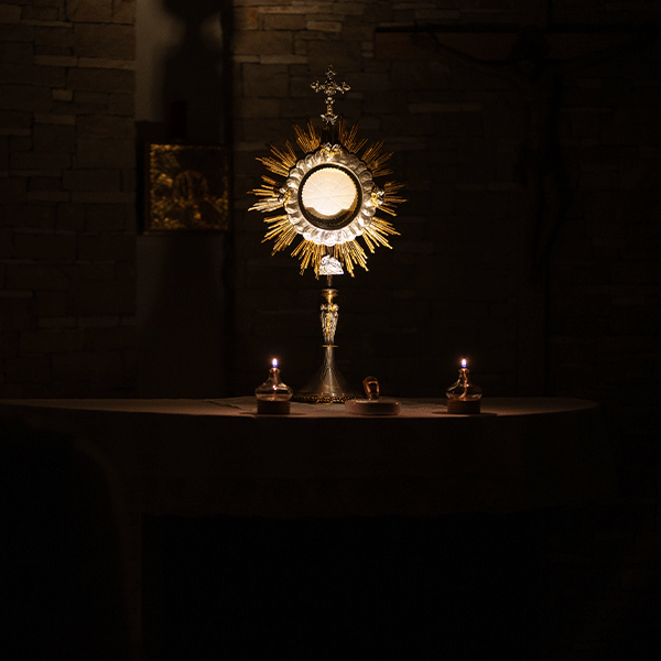 A gold monstrance containing the Eucharist sits on a table, illuminated by two small candles in a dim, stone-walled room.