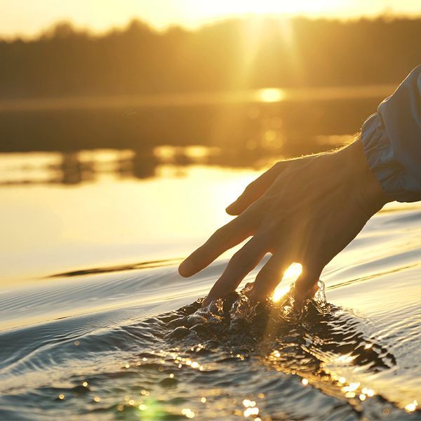 Hand touching rippling water at sunset with golden light reflecting on the surface.
