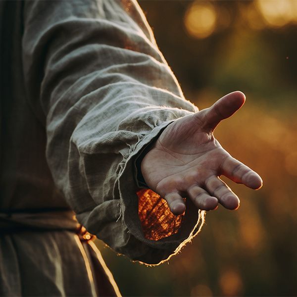 Outstretched hand in a loose beige sleeve, reaching toward warm sunset light outdoors.