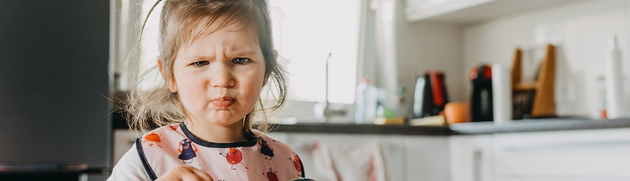 Toddler with a messy face in a kitchen, looking at the camera, wearing a light shirt and bib.